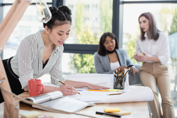 Wall Mural - Smiling young asian businesswoman working with blueprint while colleagues sitting behind