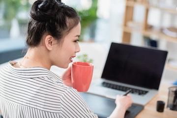 Wall Mural - young asian designer drinking coffee while working with graphics tablet and laptop