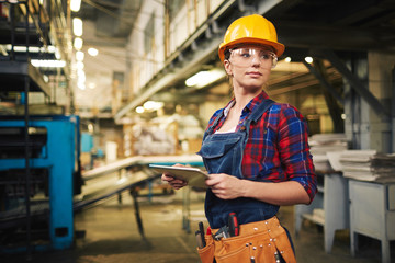 Wall Mural - Waist-up portrait of pretty young engineer looking away while working on digital tablet,  interior of manufacturing plant on background