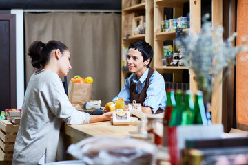 Profile view of female customer with ponytail reading label on honey jar while middle-aged shop assistant looking at her with warm smile