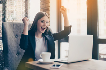 Winner! Yes! Young carefree dreamy cute girl is celebrating the breakthrough of her business, sitting in the cafe. She is confident and successful