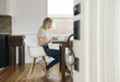 © Maskot - Full length side view of mid adult woman holding mobile phone while using laptop at home