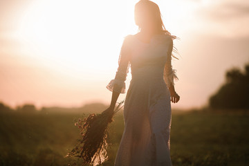 Young woman in long white lace dress on cornfield at sunset.