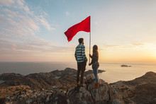Young Couple With Red Flag In A Cliff Of A Sunrise
