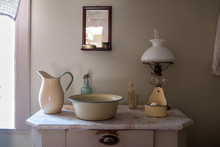 An Old Wash Basin With Water Jug, Wash Bowl, Soap Holder In Enamelled Metal. Standing On A Commode/chest Of Drawers With A Kerosene Lamp And A Mirror Above