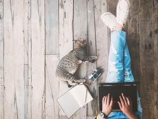 Hipster lifestyle and Creative workspace - Girl in jeans working on the laptop computer assisted by her cat on the wooden floor. vintage film color effect and retro color style