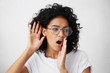 © wayhome.studio  - Curious African woman with curly hair wearing glasses and white top holding hand near her ear trying to eavesdrop interesting news. Young mixed race woman liking gossiping having intriguing look