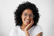 © wayhome.studio  - Close-up portrait of Afro American woman with dark curly bushy hair wearing glasses and white top holding hand on cheek looking at camera with shy charming smile feeling enjoyment and shyness