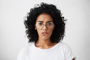 Wall Mural - Headshot of African pretty female with dark eyes, well-marked eyebrows and bushy curly hairstyle wearing big round glasses and white T-shirt biting her lip while having self-confidence and doubt