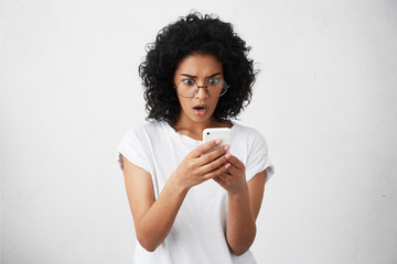 Candid shot of amazed businesswoman with bushy curly hair dressed casually holding her modern smartphone recieving message being shocked to forget about important meeting with businesspartners