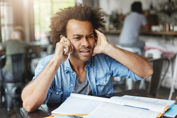 Wall Mural - Hnadsome dark-skinned college student dressed casually looking frustrated, plugging one ear while trying hard to hear what they are saying on the other end of line, asking What? Come again please?