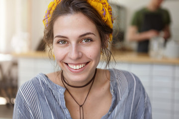 Headshot of beautiful woman with dark warm eyes pure skin and broad smile having rest at home. Caucasian female with natural beauty looking with happiness into camera while sitting in kitchen