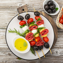 Healthy Snack: Mouth-watering Kebabs On A Picnic With Tomatoes, Mozzarella, Salami, Black Olives, Basil, Tortellini Pasta On A Cutting Board On Wooden Background With Olive Oil And Herbs. From The Top