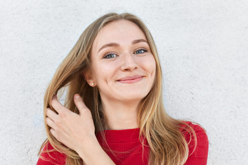 Pleased woman with blonde hair, luminous eyes and gentle smile having dimples on cheeks touching her beautiful hair with hand dressed in red clothes having good mood while posing against white wall