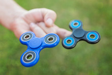 Man Holding Two Fidget Spinners On His Fingers Against The Blurred Grass Background