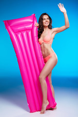 Wall Mural - Smiling young woman in bikini standing with swimming mattress and waving hand