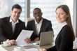 © fizkes - Attractive young businesswoman smiling for camera sitting at desk, young executive manager holding business meeting for black and white subordinates clients at background, female team lead portrait