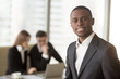 © fizkes - Attractive afro american smiling businessman looking at camera standing in foreground, young cheerful african leading manager posing with subordinates at the background, multi-ethnic team portrait