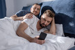 Smiling man in eyeglasses touching bearded partner using smartphone in bed