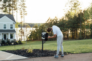 Young home owner checks on new letters and bills in his american typical mailbox, in workout clothing sweatpants on sunny warm summer evening