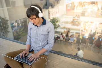 Young Asian man dressed in casual style using laptop computer while listening to music. Digital nomad working in co working space, modern IT lifestyle with work life balance concept.