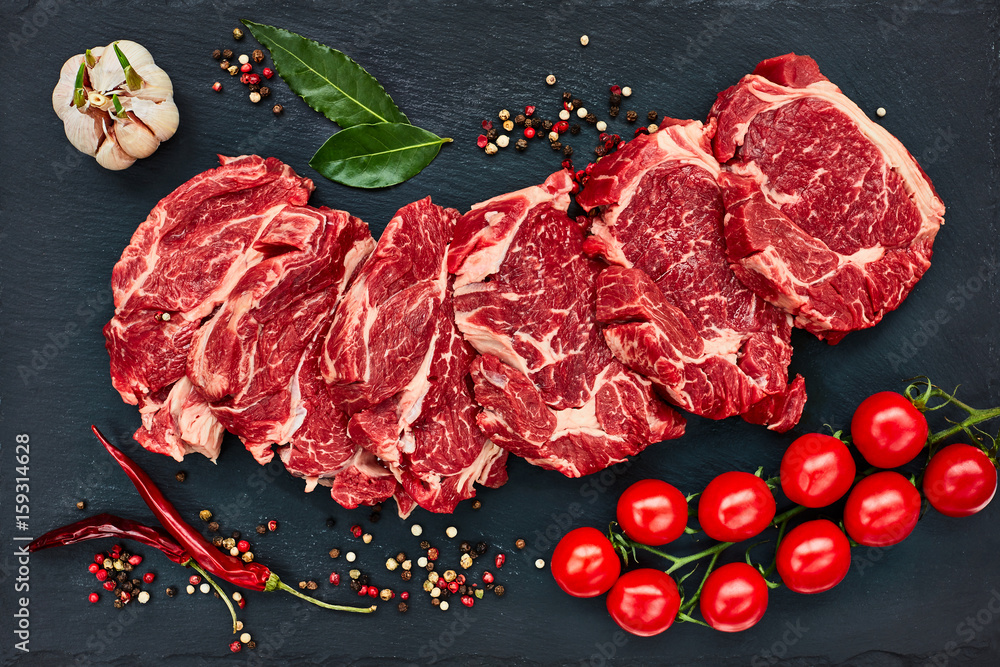 Photo Fresh raw beef steaks with pepper and tomatoes on black slate board