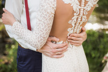 White Bridal Couple. Happy Beautiful Bride And Handsome Groom Embraces In Park Outside. Closeup Of Luxury Lacy Back Of White Dress Of Young Woman And Male Hands On Her Waist. Horizontal Color Picture.