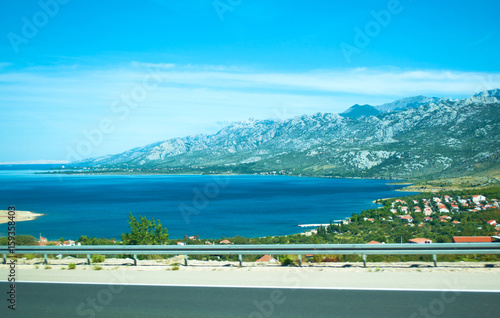 Fotografie, Obraz Highway on the background of Velebit mountain range, small houses among green trees and azure blue sea water on a summer day