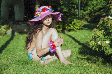 Curly young model in a hat sits on the grass and looks into the camera.