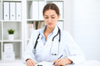 © rogerphoto - Young brunette female doctor sitting at the table and working at hospital office.