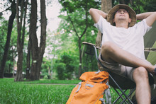 Young Man Tourist Sitting On Chair Resting And Relaxing In Front Of Tent At Camping Site In Forest. Outdoor Activity In Summer. Adventure Traveling In National Park.