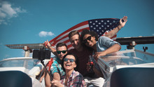 Emotional Group Of Mixed Ethnic Young Friends On Board A Motor Boat Along A River Laughing And Cheering And Raise American Flag In Air. Celebrate An Independence Day On July 4