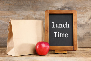 Wall Mural - Paper lunch bag, red apple and small blackboard on wooden background