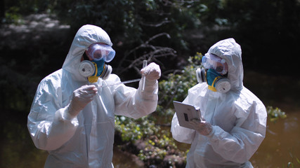 two men in biohazard suits and masks sampling water from a stream or river pipetting a sample into a