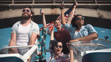 Group Of Cheering Laughing Friends On A Speedboat Waving A Patriotic American Flag As They Pass Under A Bridge In A Low Angle View Looking Up.