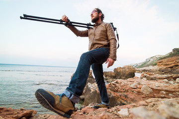 Young male photographer walking on rock beach with camera on tripod