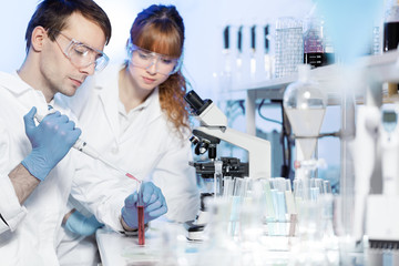 Young male researcher pipetting red liquid in the glass tube in the laboratory. Female asistant science student watching and learning the protocol.