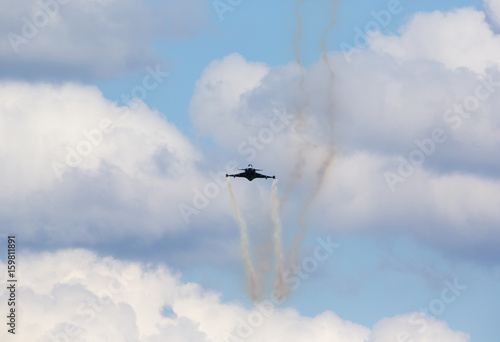 Zdjęcie XXL Myśliwiec odrzutowy latający w kierunku kamery z dymnym śladem za nim. Pochmurne niebo w tle.