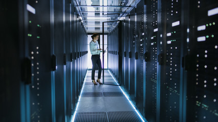 Wall Mural - Female Server Technician Stands next to Cabinet in Data Center Corridor with Rows of Rack Servers. She's Running Diagnostics on Her Computer