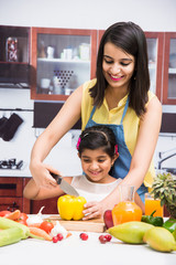 Pretty Indian young lady or mother with cute girl child or  daughter in kitchen having fun time with table full of fresh vegetables and fruits