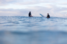 Two Out Of Focus Surfers Sit In The Distance On Their Surfboards As They Wait For A Wave To Appear On The Horizon.