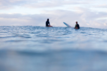 two out of focus surfers sit in the distance on their surfboards as they wait for a wave to appear o