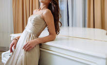 Young Beautiful Slim Brunette Girl With Long Curly Hair In Long Beige Dress With Open Shoulders Stands Near White Grand Piano In The Room