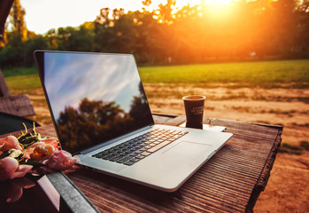 Laptop computer on rough wooden table with coffee cup and bouquet of peonies flowers in outdoor park
