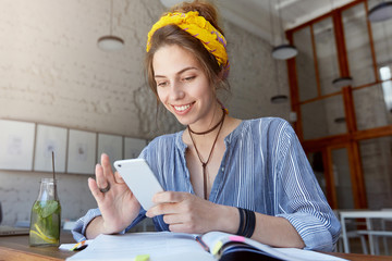 Student female wearing yellow scarf on head, stripped shirt, pendant on neck sitting in classroom surrounded with books being happy to recieve message from her friend. People, studying, technology