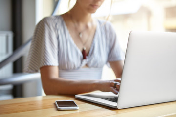 Wall Mural - Cropped view of businesswoman sitting at wooden desk indoors surrounded with modern gadgets. Clever woman working online via laptop computer typing report. Woman's hands keyboarding on computer