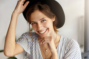 Wall Mural - Good-looking brunette woman in black hat holding her hand under chin and on hat having magic sincere smile being happy to recieve proposal from her boyfriend. Positive emotions and beauty concept