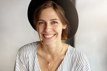 Wall Mural - Fashionable cute young lady with dark eyes, healthy skin smiling demonstrating her white teeth wearing black hat being glad to have holidays. Woman with appealing appearance rejoicing her holidays