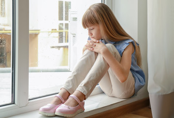 sad little girl sitting on window sill at home