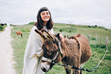 Happy And Optimistic Woman Unites With Nature By Caressing Funny Cute Donkey, Hugs Him And Laughs Into Camera